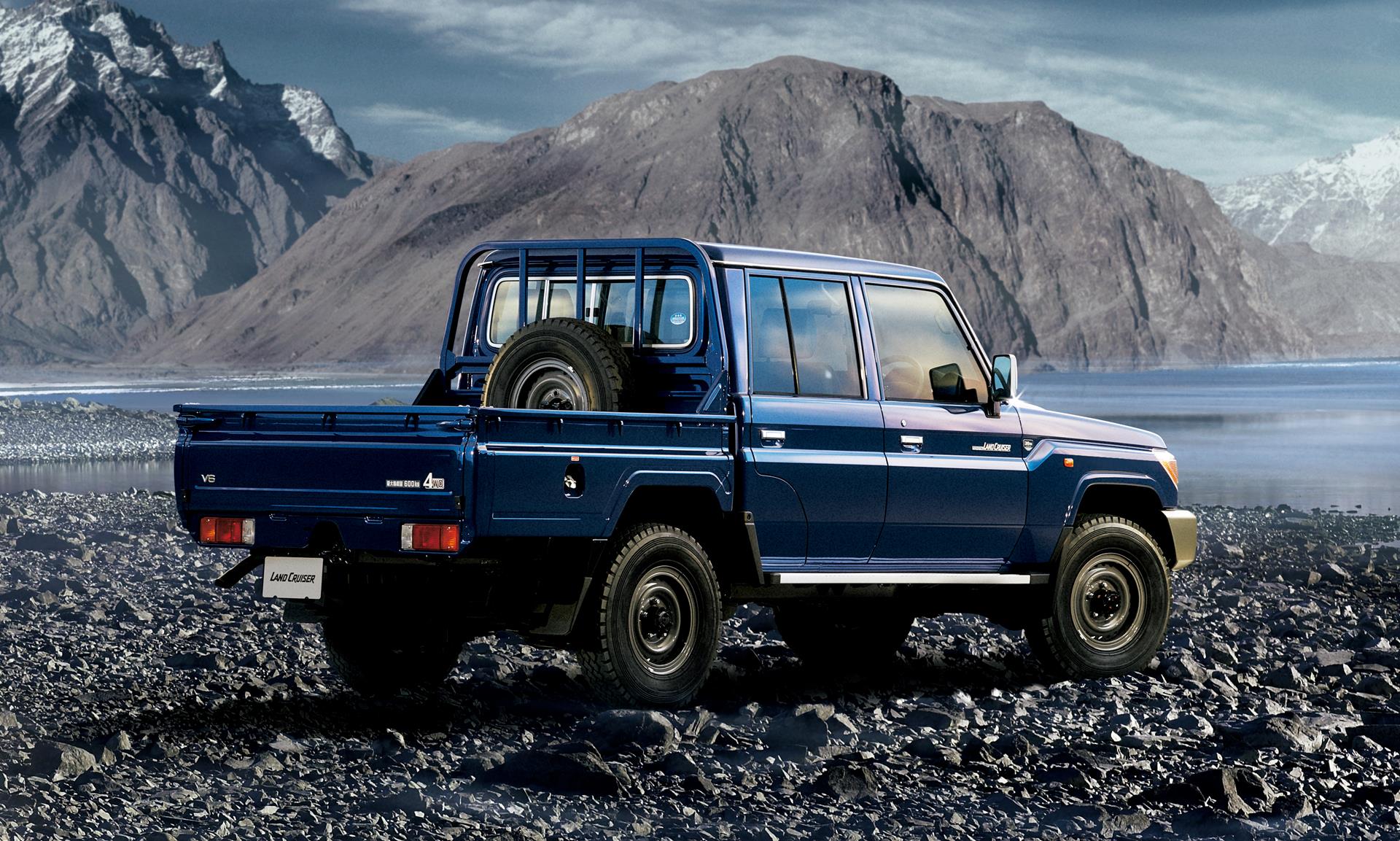 Blue Toyota Land Cruiser 70 Series double-cab pickup truck with bed-mounted spare wheel, parked on rocky ground beside a mountain lake — 15-year-plus import sourced to order in Canada